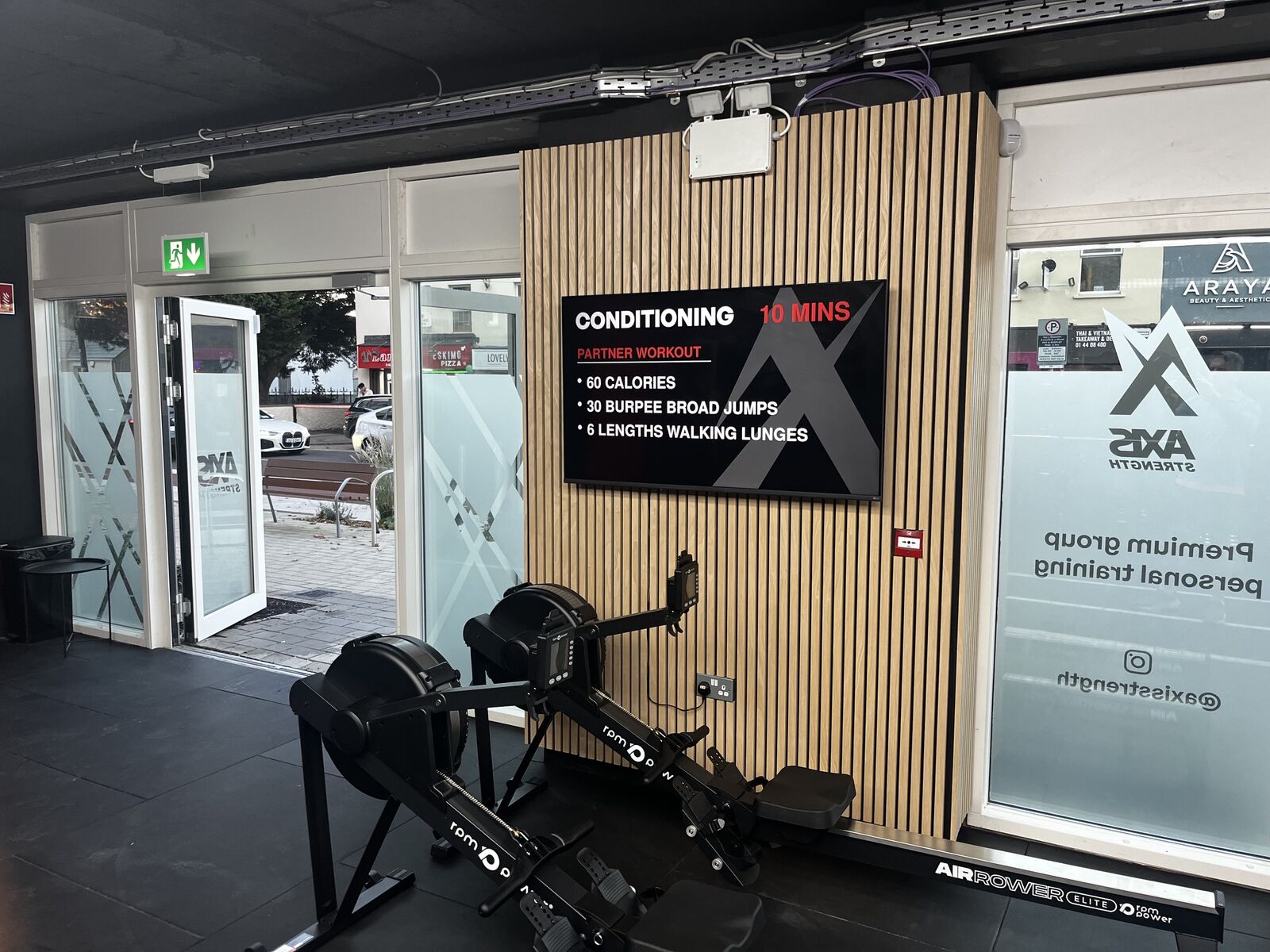 An Axis-branded conditioning workout on the studio screen, mounted on a timber slat wall above the rowing machines